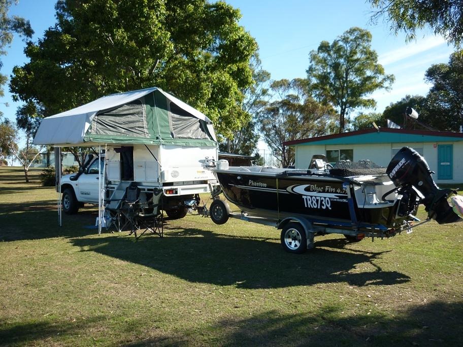A campsite with a soft top trayon slide on camper, a boat trailer, and other outdoor equipment in a grassy area surrounded by trees